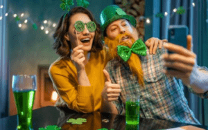 A couple sitting at a table taking a selfie with St. Patrick's day costume accessories. There are two glasses of green beer on the table in front of them.